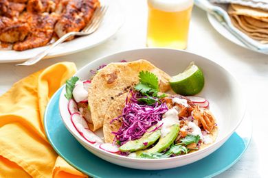 A table is set with a plate with fish tacos. Corn tortillas layered with fish, avocado, cabbage, cilantro, cheese and lime.