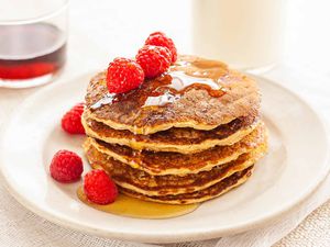 Healthy oatmeal pancakes stacked high on a whie plate drizzeled with maple syrup and topped with raspberries.