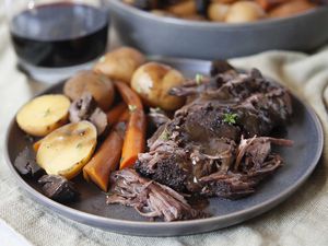 Perfectly tender crock pot roast piled onto a gray plate with carrots and potatoes and covered in gravy.