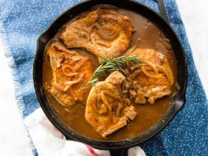 Four gluten free Smothered Pork Chops with Gravy in a skillet with a sprig of rosemary on top set against a blue background.