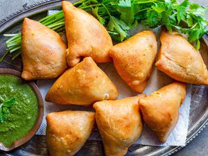 Vegetarian Samosas on a bed of herbs with one samosa filled with potato and peas alongside a cilantro mint chutney or dipping sauce in a small wooden bowl..