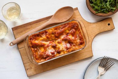Horizontal image of baked loaf pan lasagna with sausage on a wooden paddle shaped cutting board. A wooden spoon is to the left as well as two glass tumblers of white wine. A small wooden bowl of lettuce is in the upper right hand corner. In the lower right hand corner sits a stack of white plates and two forks in partial view.