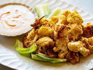 Platter of golden air fried crispy cauliflower heaped on the right of the platter. A small bowl of creamy dip swirled with hot sauce is placed next to the cauliflower on the platter. The cauliflower is resting on a few leaves of lettuce.