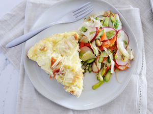 A slice of breakfast strata is on a white plate and spring salad is on the plate to the right. Sliced radish and chopped asparagus are visible in the salad. A fork is resting on the top of the plate. Cream linens are under the plate.