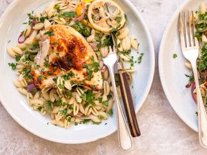 A horizontal image of two plates of yogurt marinated lemon chicken with pasta, olives and herbs. Each plate has a golden brown roasted chicken breast, roasted lemon wedges, sliced red onion, shell pasta and herbs scattered over top. The left plate has a knife and fork on the right side of the plate. The right plate is in partial view and has a fork on the left side of the plate.