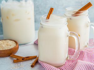 A large mason jar of easy homemade horchata with a cinnamon stick inside is in front of a similar glass and a pitcher of homemade horchata. A small bowl of rice and cinnamon sticks are to the left.