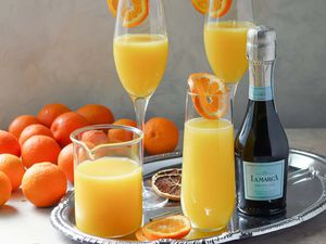 Prosecco, orange juice, orange slices and three glasses of the best mimosas are on a tray. Whole oranges are to the left of the tray.