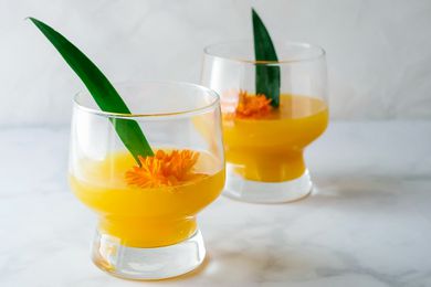 Two glasses of the best Pineapple-Mango Mimosa are on a marble counter. An orange flower and green pineapple leaf are inside each glass.