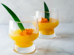 Two glasses of the best Pineapple-Mango Mimosa are on a marble counter. An orange flower and green pineapple leaf are inside each glass.