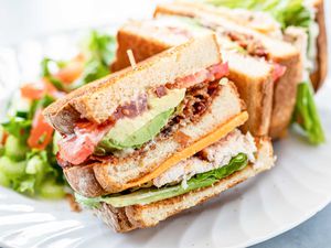 Side view of turkey club with garlic mayo on a plate. The bread is toasted and the mayo, avocado, bacon, tomato, lettuce and turkey are visible in the sandwich.