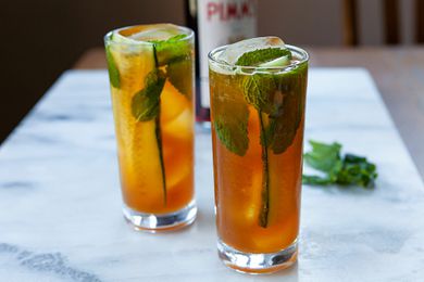 Two tall glasses of Pimms cup cocktail with slices of cucumber, mint leaves and ice. Mint leaves and Pimms no 1 are behind the glasses.