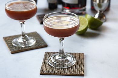 Sidecar drink with chocolate in a coupe glass with a second glass behind it. Ingredients for the cocktails are behind the glass.