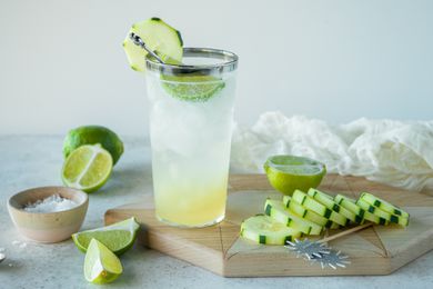 Cool cucumber lime soda garnished with cucumber and with cucumber slices and lime wedges around it.