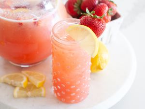 Blender strawberry lemonade served in a glass and in a pitcher with lemon slices and strawberries set around it.