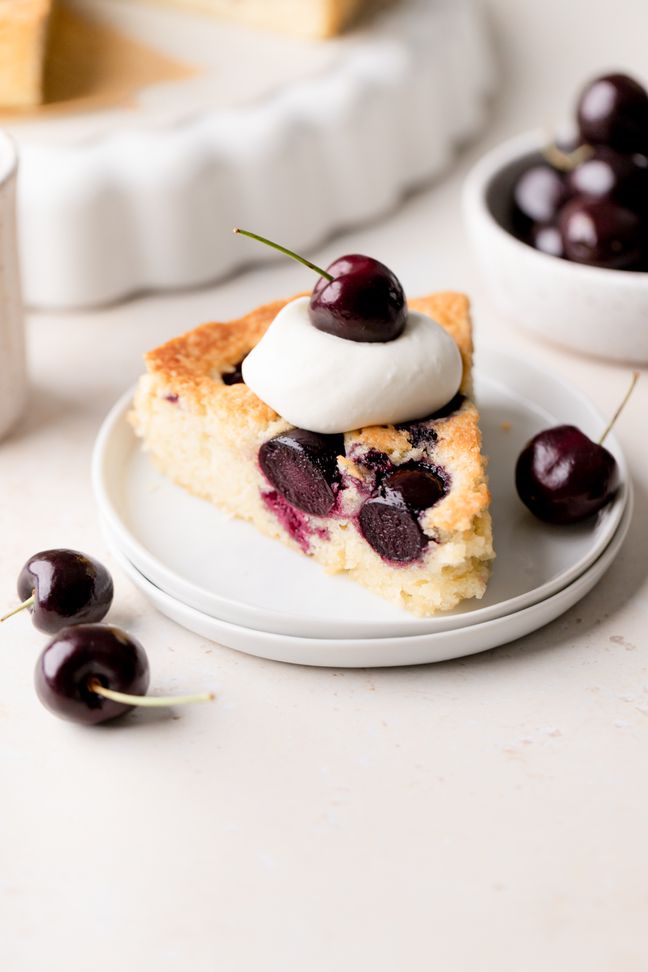 A slice of cherry cake with fresh cherries on a plate and topped with a dollop of whipped cream and a cherry.