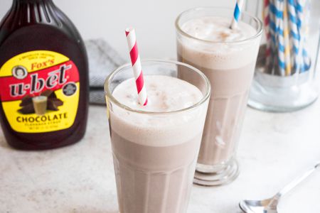 Egg Cream in Two Glasses with Bottle of Chocolate Sauce and Container of Straws in Background