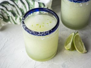Best frozen margarita in a blue rimmed glass with limes in front and another glass and linen behind it.