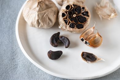Cloves of black garlic on a white plate with a whole head and a sliced head of garlic behind it.