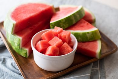 Bowl of watermelon cubes with watermelon slices behind it on a wood serving platter