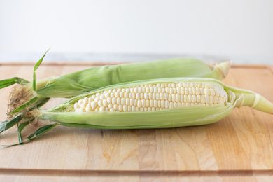 Two ears of corn with husk pulled back showing kernals on a wood cutting board