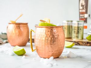 Mezcal Mule in Two Copper Mugs Surrounded by Ice and Limes. More Ingredients in the Background