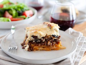 A piece of eggplant moussaka on a plate with a glass of wine and a salad behind it.