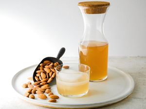 Homemade orgeat in a pitcher behind a cocktail and a scoop with almonds.