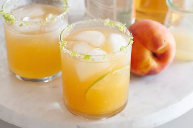 Two summery peach margaritas on a white tray with a whole peach next to it.