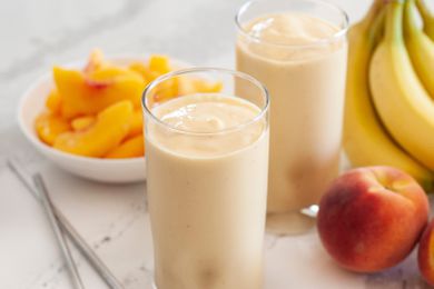 Two peach almond smoothies with whole bananas, peaches, and a plate of sliced peaches set around the smoothies.