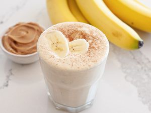 Simple peanut butter banana smoothie topped with sliced bananas and a bunch of bananas and a small container of peanut butter behind it.