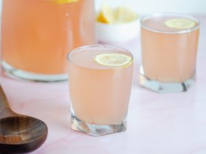 Two glasses of pink lemonade garnished with lemon rounds. A wooden spoon and pitcher are next to the glasses.