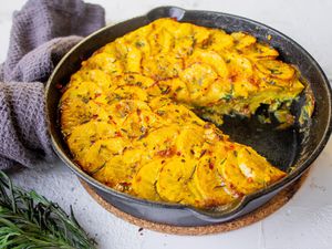 Plantain Frittata with a Serving Cut Out in Cast Iron Skillet Surrounded by Rosemary and Kitchen Towel