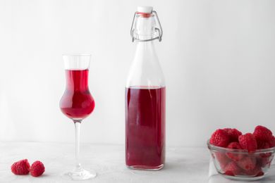 A flip top bottle of fresh raspberry liqueur with a stemmed glass and a bowl of raspberries next to it.