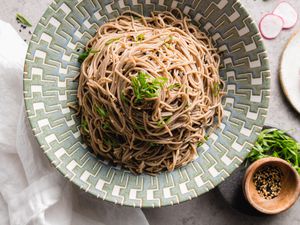 Sesame Soba Noodles in a Bowl Garnished with Spring Onions Next to Plate with Spring Onions and Seasoning and Another Plate with Slices of Radish